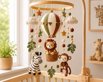May include: A crochet baby mobile featuring a central hot air balloon with a lion in the basket, flanked by a zebra and a monkey. The mobile includes hanging elements like clouds, stars, and leaves in shades of green, brown, and cream.
