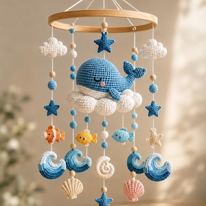 May include: A hanging mobile featuring crocheted figures suspended from a circular wooden frame. The elements include a blue whale on a white cloud, white clouds, blue and beige starfish, small orange, yellow, and blue fish, blue waves, and seashells.