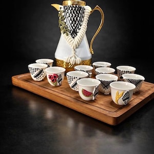 May include: A white and gold ceramic coffee pot with a decorative rope and black and white patterned accents. The set includes small white cups with various fruit and geometric designs, arranged on a wooden tray.