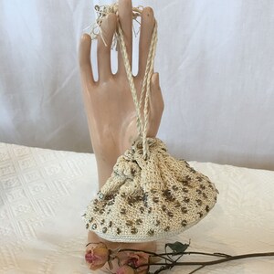 May include: A white crocheted drawstring bag with gold beads. The bag is hanging from a hand-shaped display stand.