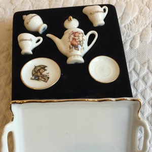 May include: A miniature tea set with a teapot, creamer, sugar bowl, two cups and saucers, and a tray. The set is decorated with a floral pattern and a small girl with an umbrella. The set is displayed on a black velvet background.