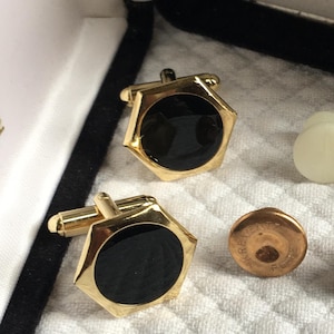May include: A set of gold-toned cufflinks and shirt studs. The cufflinks have a hexagonal shape with a black center. Two white shirt studs and two copper-colored studs are also included, all displayed on a white, textured surface.
