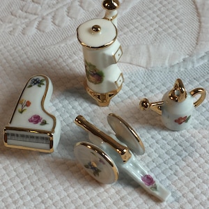 May include: A collection of miniature porcelain figurines, including a piano, a cannon, a watering can, and a decorative object. Each piece is white with gold trim and floral accents. The piano has a black and white keyboard.