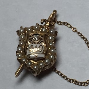 May include: A gold-toned shield-shaped pin featuring a central white enamel scroll with Greek letters, a small clear stone, and a border of small white pearls. A gold sword extends vertically behind the shield, connected to a delicate gold chain.