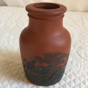 Puede incluir: Un jarrón de terracota con un diseño verde y marrón de una escena de caza. El jarrón tiene un cuerpo redondeado y una boca ancha.