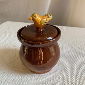 May include: A brown ceramic jar with a lid featuring a yellow bird figurine. The jar has a rounded shape and a glossy finish.