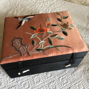 以下が含まれることがあります： 長方形の黒い箱で、桃色の蓋が付いています。 蓋には、青、オレンジ、緑、茶色の鳥、花、葉が刺繍されています。 箱には留め金が付いています。