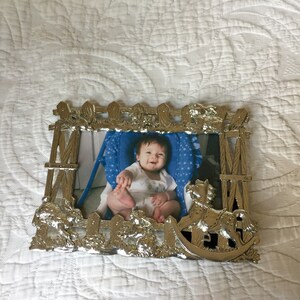 Pode incluir: Moldura de fotos prateada com um design de cerca branca. A moldura apresenta um cavalo de balanço e uma foto de um bebê em uma cadeira alta azul e branca.