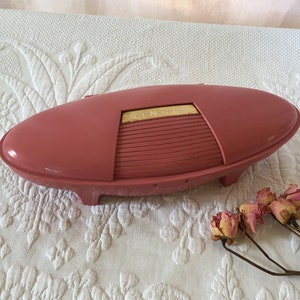 Puede incluir: Un estuche de máquina de coser Singer vintage de color rosa con una etiqueta dorada. El estuche tiene forma de barco y presenta un diseño acanalado en la parte superior.