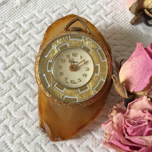May include: A gold-toned wristwatch with a white face and black Roman numeral hour markers. The watch is set in a gold-toned metal case and is attached to a brown agate pendant.
