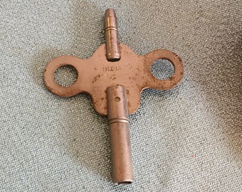 May include: A vintage metal clock key with a  "INDIA" inscription. The key has a long, thin shaft and a three-pronged head.
