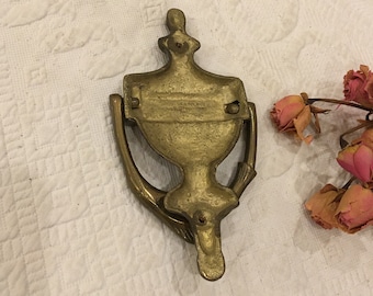 Könnte beinhalten: Ein Türklopfer aus Messing in Form einer stilisierten Urne, die von zwei Händen gehalten wird. Der Türklopfer hat ein dekoratives Design und ist in gutem Zustand.