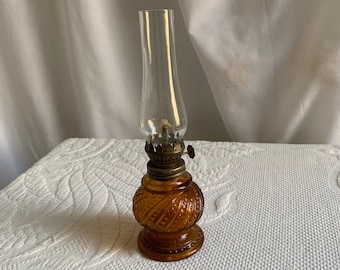 May include: A small amber glass oil lamp with a clear glass chimney and a brass wick adjuster. The lamp has a textured, swirl design.