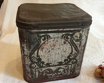 Könnte beinhalten: Vintage Metalldose mit quadratischer Form und einem Scharnierdeckel. Die Dose ist mit einem weißen Hintergrund und einem schwarzen und roten Blumendesign verziert. Die Dose weist Gebrauchsspuren auf, mit Rost und Kratzern auf der Oberfläche.