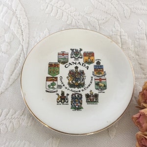 May include: A white ceramic plate with a gold rim features the coats of arms and emblems of the Canadian provinces and territories. The plate is decorated with colorful illustrations of each emblem, including the Canadian coat of arms in the center. The text "CANADA" and "COATS-OF-ARMS & EMBLEMS" are printed in black.
