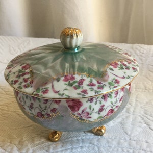 Może przedstawiać: Porcelanowa cukiernica z pokrywką i trzema nóżkami. Miska jest ozdobiona kwiatowym wzorem w odcieniach różowego, zielonego i złotego.