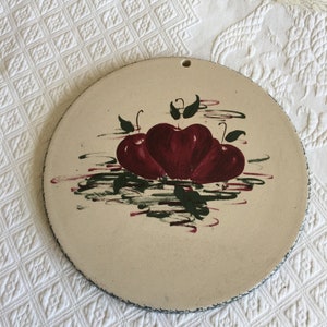 以下が含まれることがあります： クリーム色の背景に、緑の葉のついた赤いリンゴ3つが描かれた、丸いセラミック製の鍋敷きです。