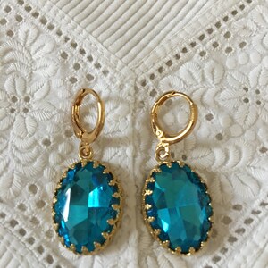 May include: A pair of gold hoop earrings with a large, oval-shaped, teal blue gemstone. The gemstone is faceted and has a gold setting.
