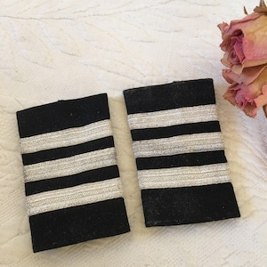 Puede incluir: Dos charreteras de tela negra con tres rayas plateadas cada una. Las charreteras son para un uniforme.