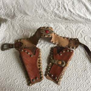 Op de afbeelding: Een bruine leren speelgoedpistoolholster met zilveren studs en een rood juweel. De holster heeft twee holsters en een riem met een zilveren gesp.