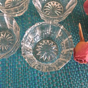 May include: A collection of clear, faceted glass bowls with a starburst design in the center. The bowls are hexagonal in shape and sit on a teal and blue woven surface. A dried rose is visible in the background.