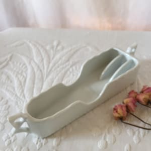 Puede incluir: Una fuente de cerámica blanca con una forma alargada única y dos divisores internos. La fuente tiene un borde ondulado y pequeñas asas en cada extremo. Rosas rosas secas en el fondo.