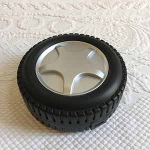 Puede incluir: Un contenedor negro con forma de neumático con un diseño de estrella plateada en la tapa. El contenedor está hecho de plástico y tiene una superficie texturizada.