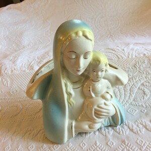 May include: A ceramic planter shaped like the Virgin Mary holding the baby Jesus. The planter is painted in shades of blue, white, and yellow.