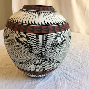 Puede incluir: Un jarrón redondo de cerámica blanca con diseños geométricos negros y rojos. El jarrón tiene una base ancha y un cuello estrecho.