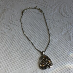 May include: A silver-toned chain necklace featuring a triangular pendant with rounded corners. The pendant displays an intricate openwork scroll design in silver metal, accented with small, smooth, circular gold-toned metal beads.