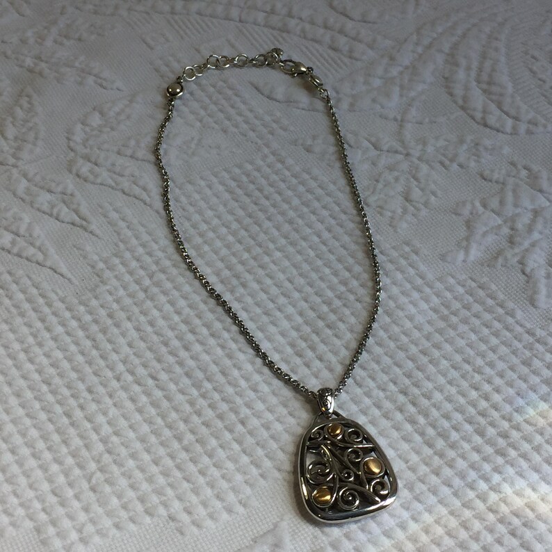May include: A silver-toned chain necklace featuring a triangular pendant with rounded corners. The pendant displays an intricate openwork scroll design in silver metal, accented with small, smooth, circular gold-toned metal beads.
