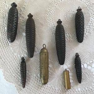 May include: An assortment of dark brown, cone-shaped clock weights and two cylindrical weights. The weights vary in size and have small metal loops at the top. The background is a white, lacy fabric.