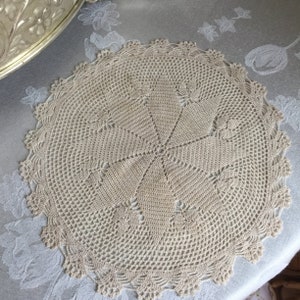 以下が含まれることがあります： 円形のデザインと繊細なレースの縁取りが施されたベージュのクロシェのドイリーです。