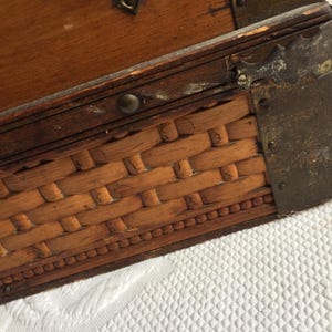 Puede incluir: Un cofre de madera con un fondo de mimbre tejido y detalles met&aacute;licos. El cofre tiene un tinte marr&oacute;n oscuro y muestra signos de desgaste.