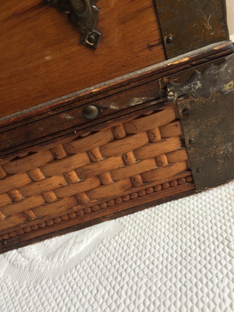 Puede incluir: Un cofre de madera con un fondo de mimbre tejido y detalles met&aacute;licos. El cofre tiene un tinte marr&oacute;n oscuro y muestra signos de desgaste.