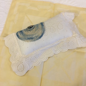 Könnte beinhalten: Ein weißes Kissen mit einem blauen und weißen gehäkelten Kreis oben. Das Kissen hat eine weiße Spitzenborte und liegt auf einem gelben Stoff.