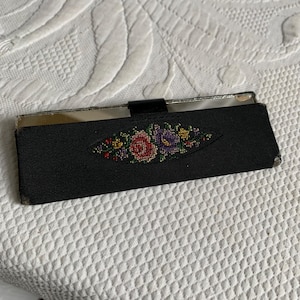 以下が含まれることがあります： ピンク、パープル、グリーンの色の花柄刺繍が施された黒い布製のコンパクトミラー。ミラーはシルバーフレームで、黒い革の留め具が付いています。