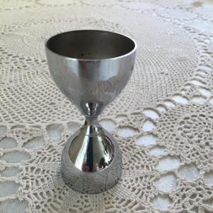 Könnte beinhalten: Ein glänzender, silberfarbener Cocktail-Jigger mit ausgestellter Oberseite und rundem Boden. Der Jigger hat einen schmalen Stiel, der die beiden Teile verbindet. Die Oberfläche reflektiert die Umgebung. Der Jigger wird zum Messen von Flüssigkeiten für Cocktails verwendet.