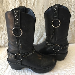 Könnte beinhalten: Ein Paar schwarze Leder-Cowgirl-Stiefel mit silbernen Nieten und Schnallen. Die Stiefel haben einen Absatzblock und eine spitze Zehenpartie.
