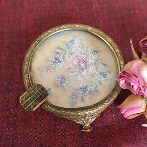 Könnte beinhalten: Eine runde, goldfarbene Schmuckschatulle mit einem floralen Design auf dem Deckel. Der Deckel ist aus Glas und das Design ist mit rosa und blauen Fäden bestickt. Die Schachtel hat einen kleinen, dekorativen Griff an der Unterseite.