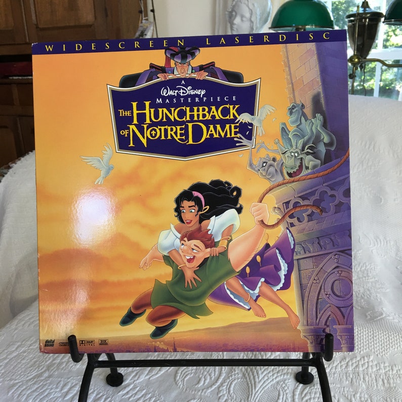 May include: A vintage laserdisc case for the Disney animated film "The Hunchback of Notre Dame". The cover features a colorful illustration of the characters Quasimodo, Esmeralda, and Phoebus flying through the air. The text "A Walt Disney Masterpiece" is printed in a gold font.
