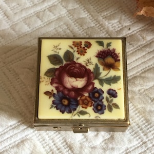 Pode incluir: Uma pequena caixa de joias quadrada de latão com uma tampa articulada. A tampa apresenta um desenho floral com uma grande rosa vermelha e flores azuis e amarelas menores sobre um fundo creme.