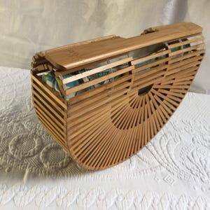May include: A light brown, semi-circular bamboo handbag with a hinged lid. The bag's body is constructed of thin, parallel bamboo slats, fanning out from the bottom. The interior lining is visible, featuring a colorful floral pattern. The bag is open.