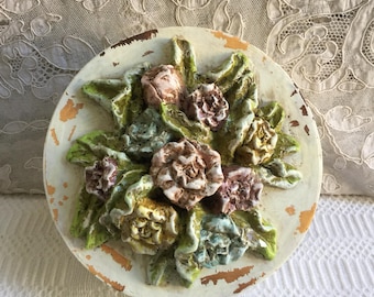 May include: A round, off-white ceramic wall plaque featuring a raised floral design. The flowers are in shades of pink, yellow, and blue, with green leaves. The plaque has a distressed, aged appearance with visible paint chipping.