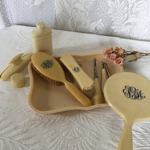 May include: An antique vanity set in cream color, including a brush, mirror, tray, and other accessories. Each piece is adorned with a monogram in dark blue. The set is arranged on a white lace surface, with dried flowers.