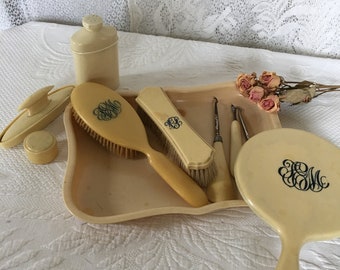 Può includere: Un set da toeletta antico color crema, composto da spazzola, specchio, vassoio e altri accessori. Ogni pezzo è decorato con un monogramma blu scuro. Il set è disposto su una superficie di pizzo bianco, con fiori secchi.