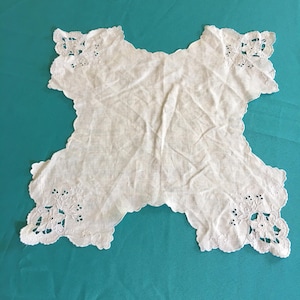 May include: A white, scalloped-edge doily with embroidered floral details at each corner. The doily has a unique shape with four extended points and a delicate, openwork design. The fabric appears to be a lightweight linen or cotton.