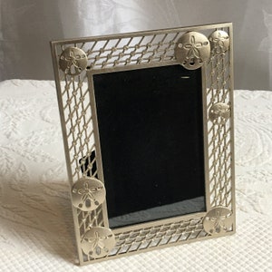 Puede incluir: Un marco de fotos plateado con un paspartú negro y un diseño decorativo de conchas alrededor del borde.