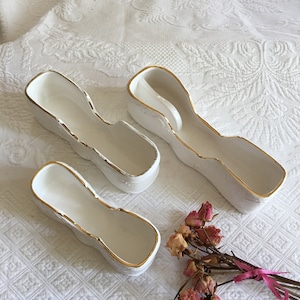 Puede incluir: Tres platos de cerámica blanca con ribete dorado. Los platos tienen forma de zapato y tienen una pequeña hendidura en el centro. Los platos están sobre una superficie blanca.