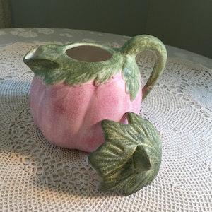 May include: A ceramic pitcher with a pink body and green leaf-shaped accents. The pitcher has a handle and a spout, and the lid is off to the side. The pitcher is designed to look like a fruit.